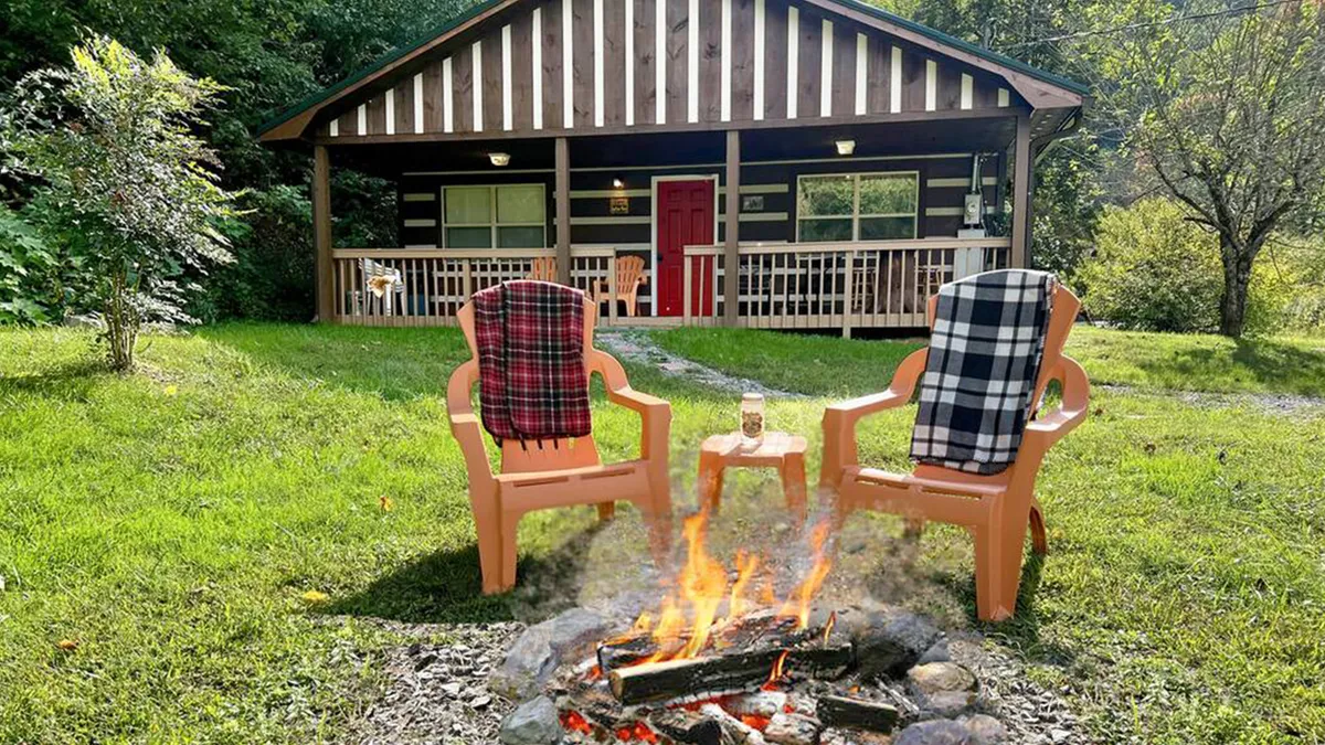 The image showcases the cabin's charming exterior surrounded by lush greenery, with a welcoming fire pit encircled by wooden chairs in the spacious yard. The inviting front porch hints at relaxation, while the fire pit setup suggests evenings spent gathering under the stars.