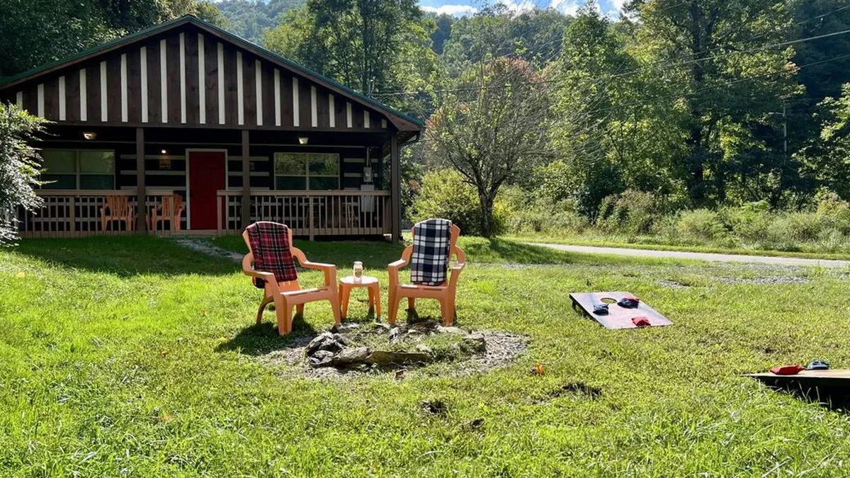 This image captures the cabin's exterior with the fire pit, lawn chairs, and a visible cornhole game set up in the spacious, flat front yard. The atmosphere is casual and communal, showing both the play and relaxation aspects of the outdoor space.