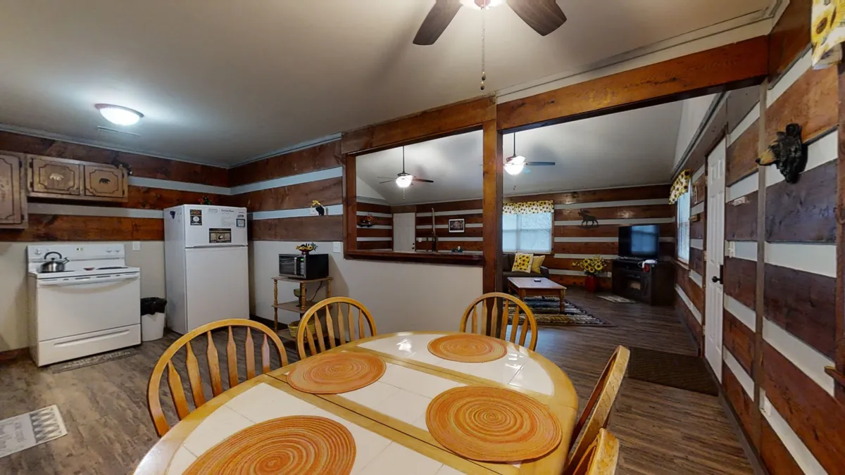 This photo showcases a spacious dining table with orange circular placemats, surrounded by wooden cabinetry and with a clear view into the adjacent living area. The table is set for a group, suggesting family meals or gatherings, while the kitchen and living room are both visible, emphasizing the open-plan layout.