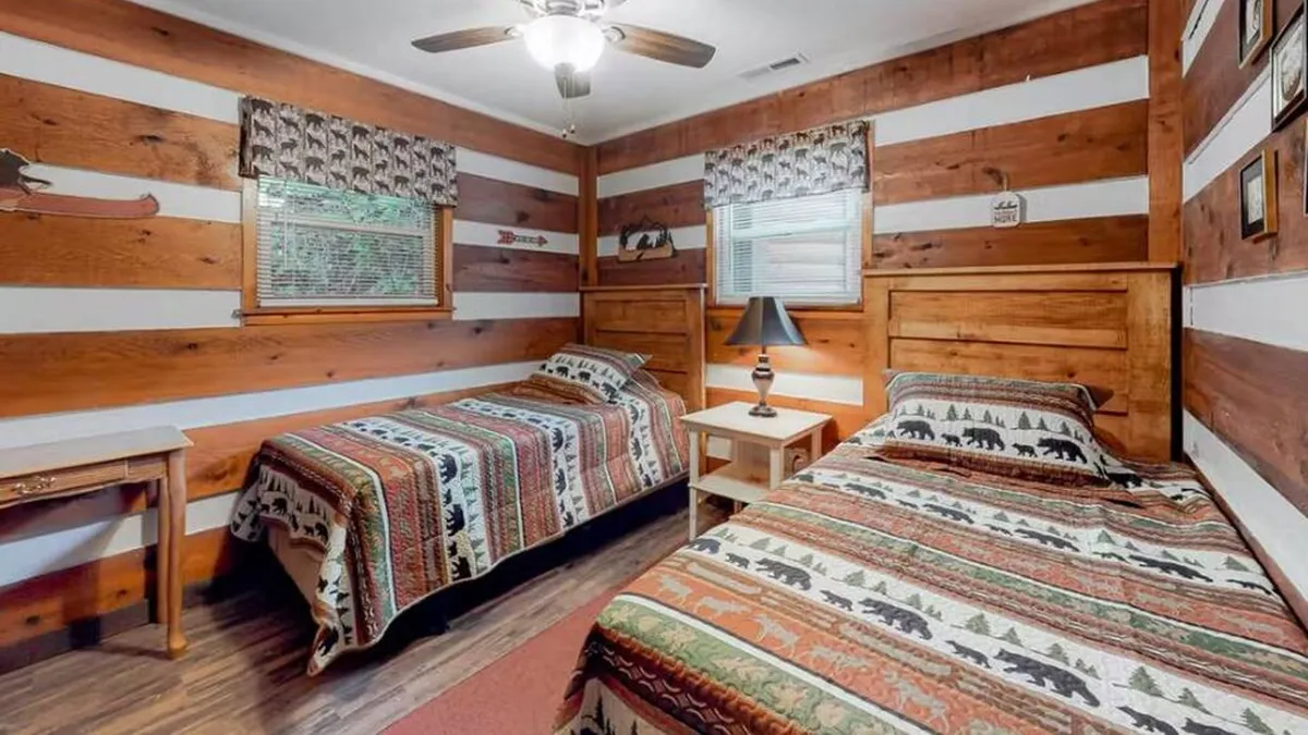 A warmly lit bedroom with two twin beds, decorative bedding, a nightstand with a lamp, and wooden walls. The space feels welcoming and comfortable, ideal for children, friends, or extra family members to unwind and share stories.