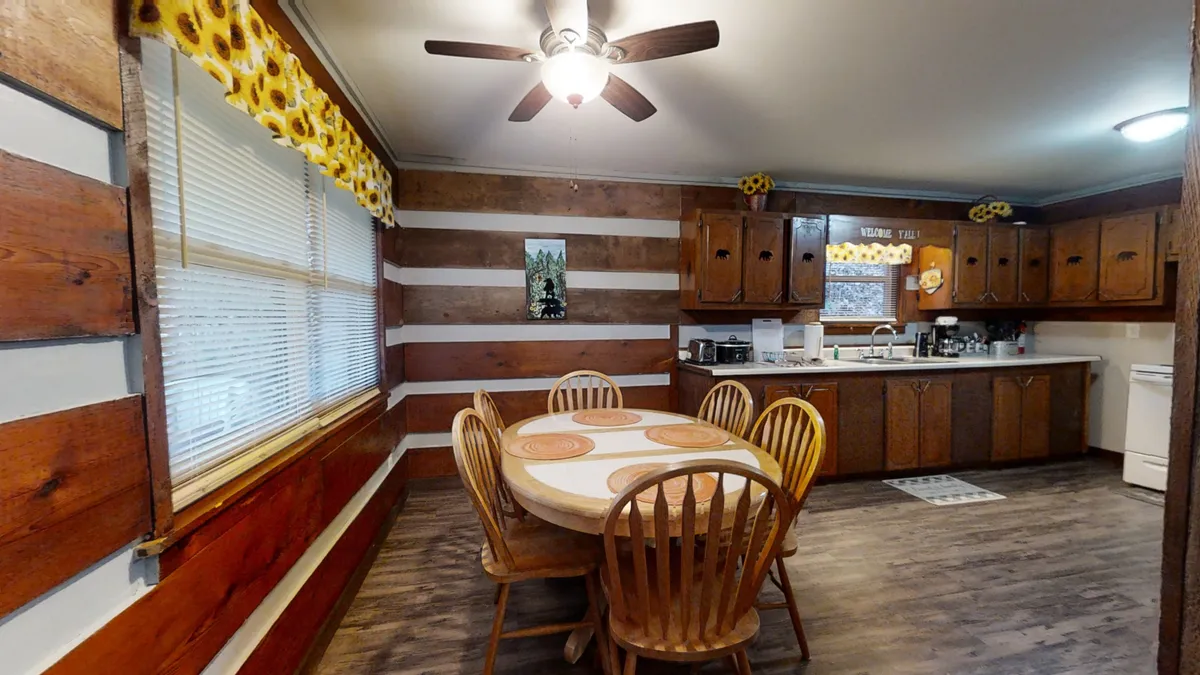 A warm, inviting dining area with a round wooden table, surrounded by matching chairs and wooden cabinetry. Sunflower-themed curtains brighten the setting, and a window lets in soft natural light. The space is perfect for family meals, casual gatherings, or board games.