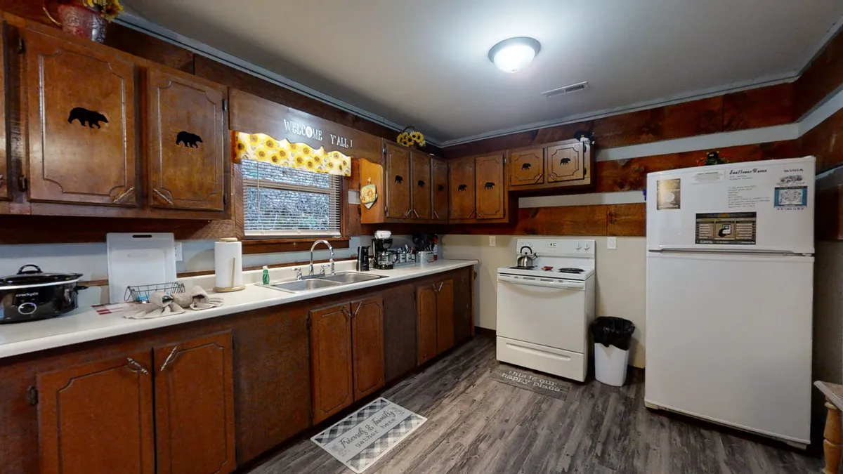A homely kitchen scene with wooden cabinetry adorned with bear motifs, a spacious central countertop, white appliances, and a window dressed in floral curtains. The space feels inviting and is ideal for enjoying a fresh cup of coffee or preparing a meal before or after exploring the valley.