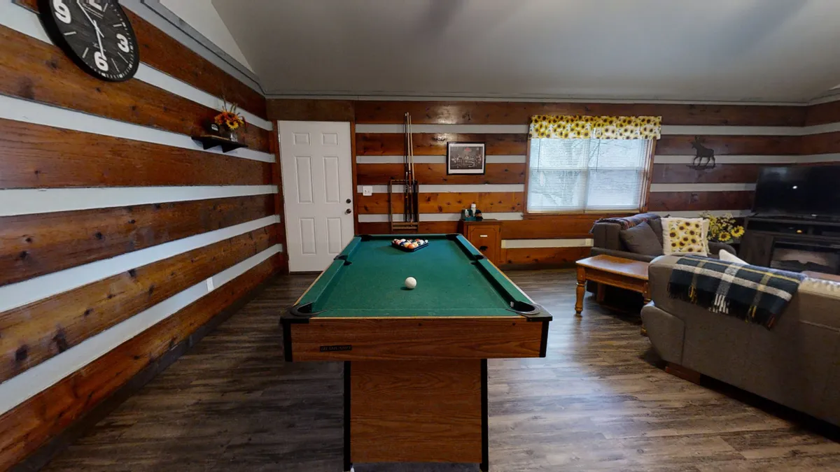 A spacious living area featuring a pool table, plush sofa, decorative wall elements, a large window letting in natural light, and a visible clock. The room is casual with areas for both relaxation and entertainment.
