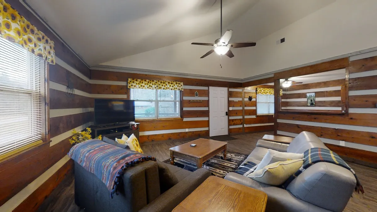 This image features a warm, welcoming living space with log walls, comfortable seating, a central coffee table, and expansive window views. The area exudes a cozy mountain cabin ambiance, perfect for relaxing after a day of exploring or enjoying a morning cup of coffee as sunlight pours in.