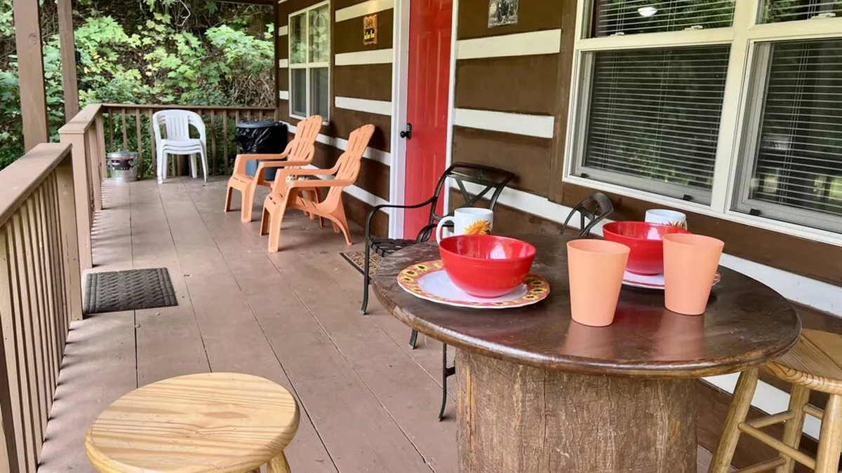 This exterior image features a welcoming porch with a wooden table set with colorful dishes and orange chairs, set against a red door. The scene exudes a casual, friendly vibe, inviting families to gather and enjoy outdoor dining as the sun sets, with beautiful views and the sounds of nature in the background.